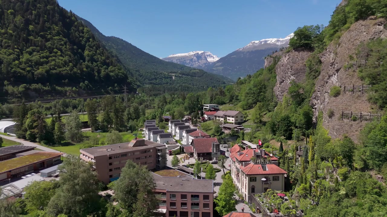 Flygfoto med drönare över Rothenbrunnen, Schweiz, som visar en alpby omgiven av gröna kullar, skogsklädda sluttningar och berglandskap i starkt dagsljus.