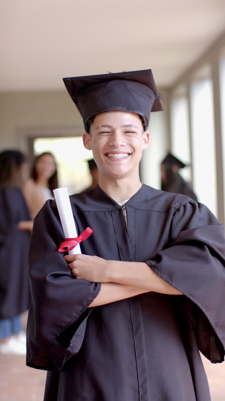 Vertical video: Teenager in graduation gown holding diploma, smiling proudly in school hallway