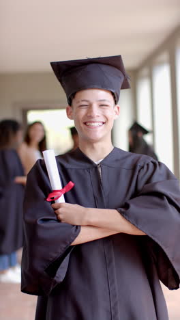 vertical video: teenager in graduation gown holding diploma, smiling proudly in school hallway