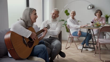 happy senior woman singing and playing the guitar sitting on chair, while in blurred background elderly friends listening to her and singing together sitting at the table 1