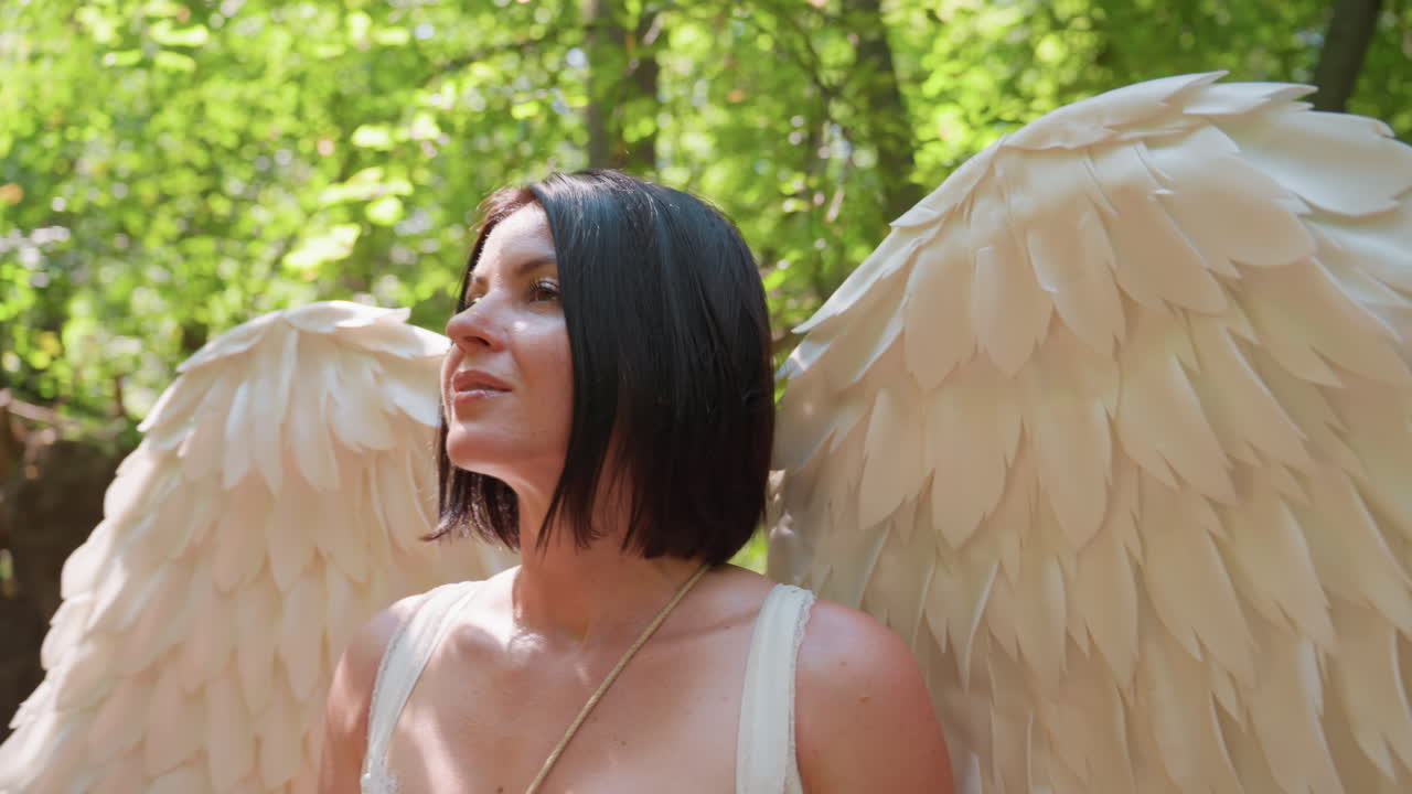 Fairy with large white wings and wooden staff gazes upward surrounded by green forest trees under warm sunlight, expressing calm wonder and spiritual connection