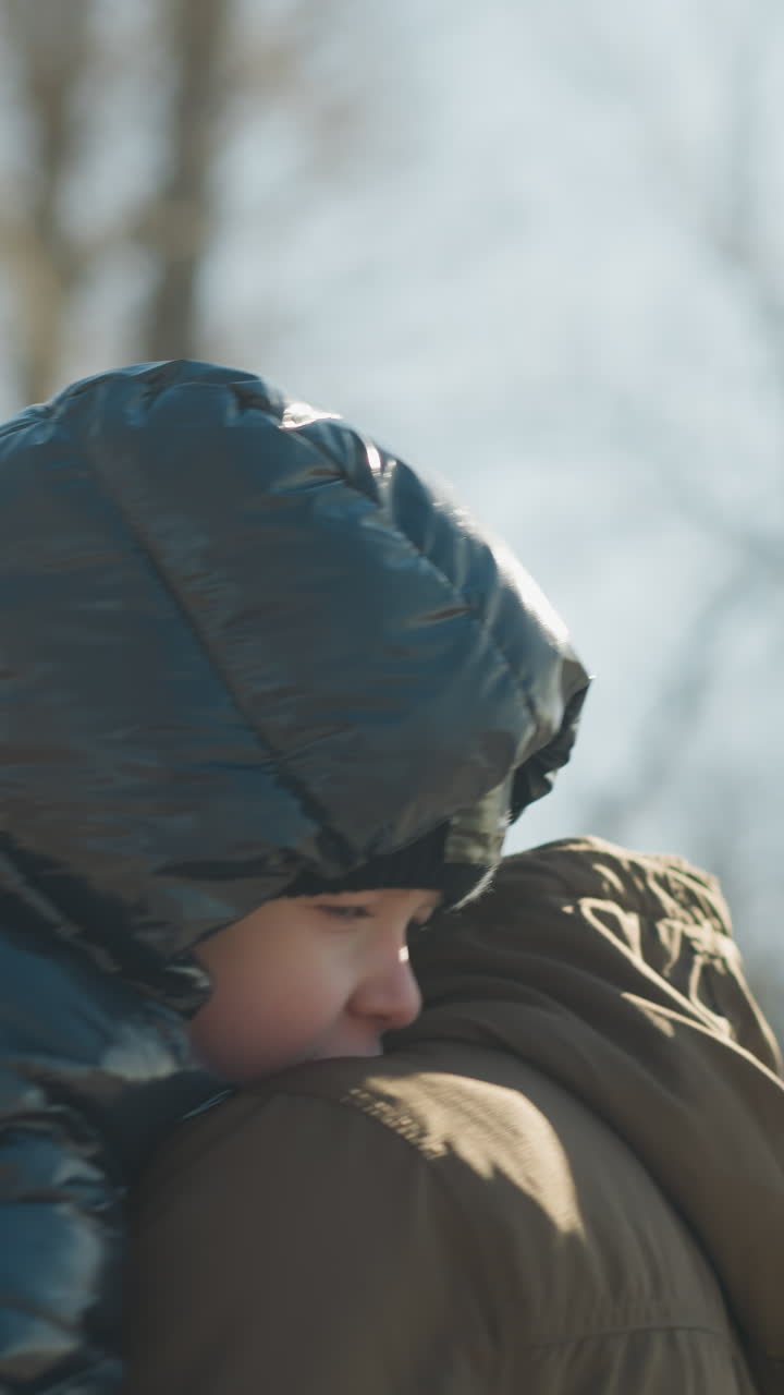 는 동안 아이를 들고 있는 아버지의 뒷면, 아버지는 갈색 재과 베니를 입고 있으며, 아이는 검은색 부풀어오르는 재을 입고 있으며 나무와 하늘이 흐릿합니다.