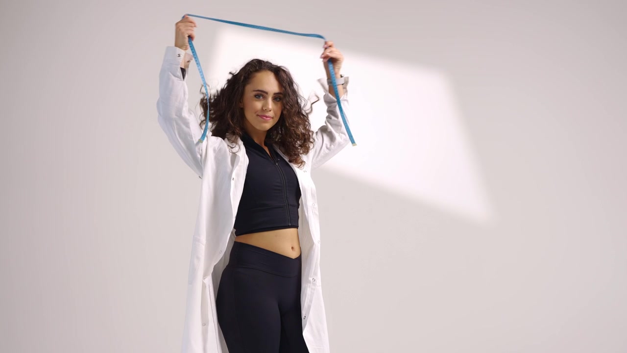 Junge lächelnde Sport-Ernährungsberaterin in schwarzer Sportkleidung und weißem Arztkittel hält ein Maßband vor weißem Hintergrund. Konzept für gesunden Lebensstil, Fitness, Ernährung und Wohlbefinden.