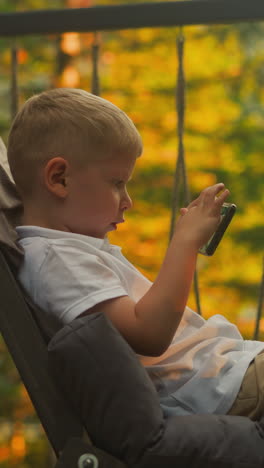 serious boy holds cellphone sitting alone on balcony at sunset. focused toddler kid plays video games enjoying atmospheric location with incredible view in background