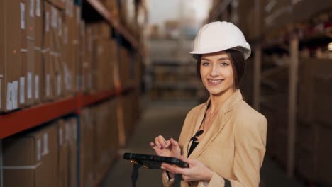 retrato de una hermosa joven en un traje de negocios con un casco en la cabeza trabajando con una tableta en sus manos en el fondo del almacén con cajas