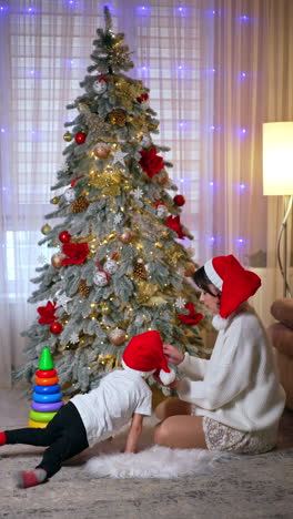brunette woman in white sweater and red cap pulls a pyramid toy. little toddler boy jumps up and sits down beside. beautiful christmas tree at backdrop. vertical video.