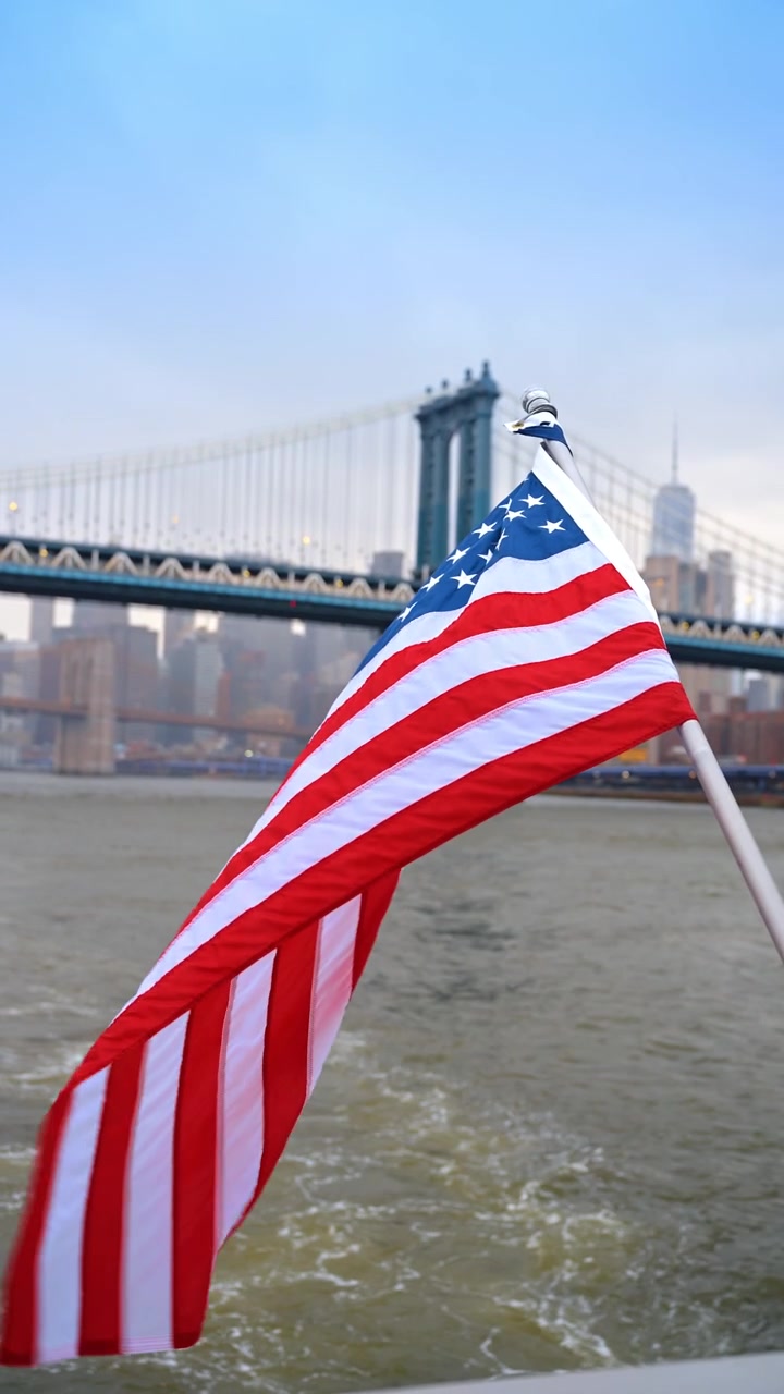 Vista dal traghetto della bandiera americana con il Ponte di Brooklyn, New York. Sullo sfondo, il ponte e i grattacieli sfocati dietro una bandiera nitida che sventola su un fiume calmo.