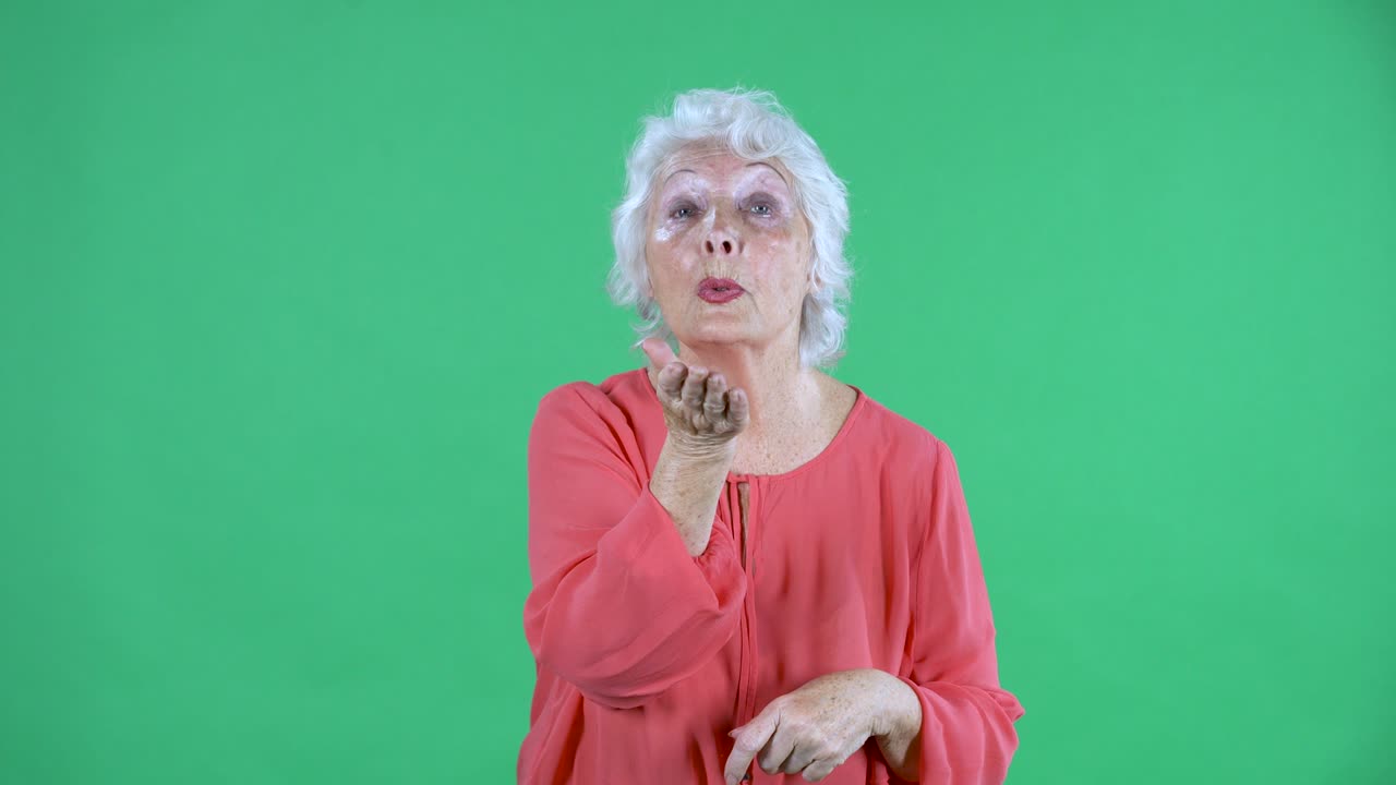 Portrait elderly woman looking at the camera and shows a heart gesture with her hands and sends an air kiss. Gray haired grandmother in red blouse on green screen at studio. Close up