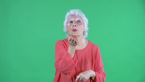portrait elderly woman looking at the camera and shows a heart gesture with her hands and sends an air kiss. gray haired grandmother in red blouse on green screen at studio. close up