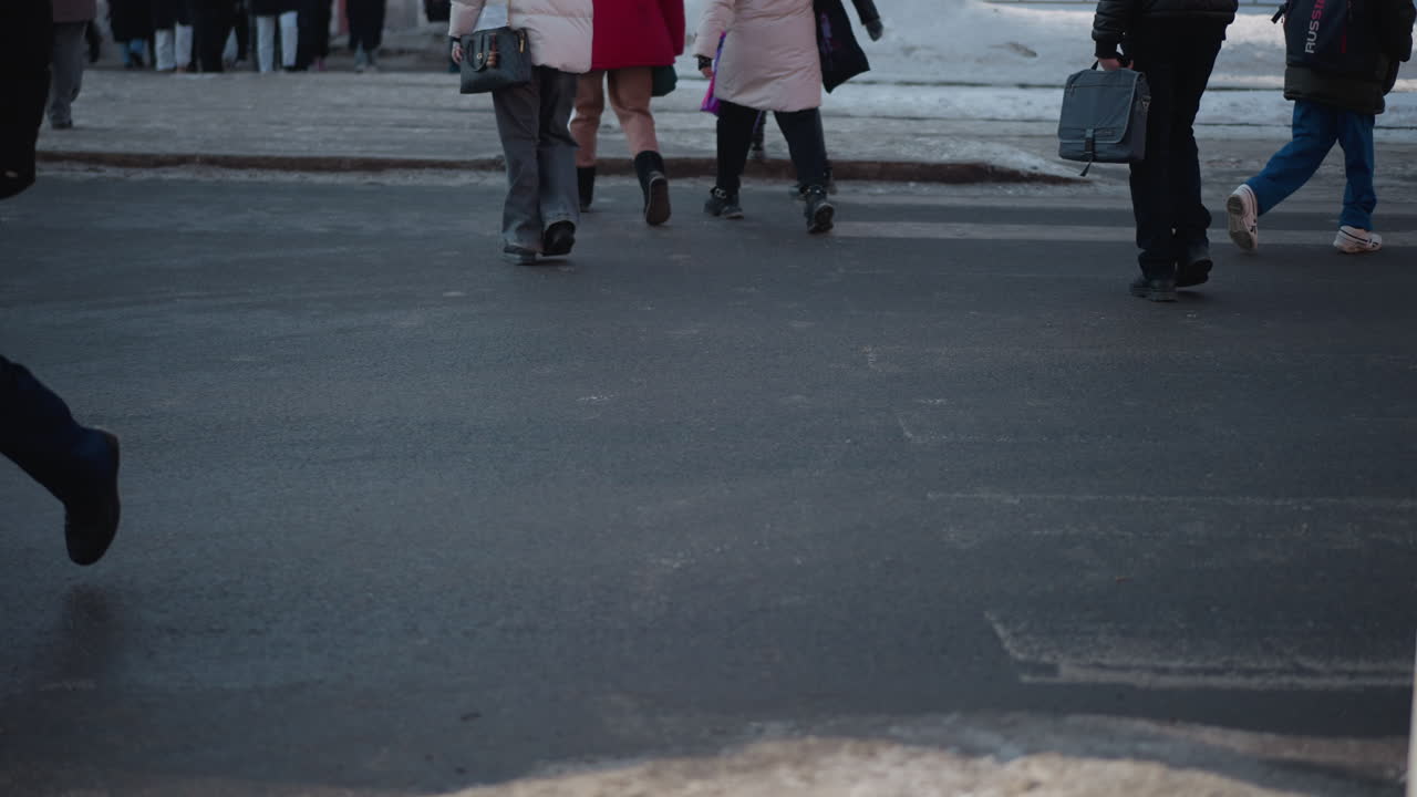 Pedoni di spalle in cappotti invernali che camminano oltre un palo segnaletico su una strada cittadina, con una borsa, che scendono dal marciapiede fangoso sull'asfalto bagnato sotto il sole, movimento verso l'attraversamento pedonale, freddo tragitto urbano