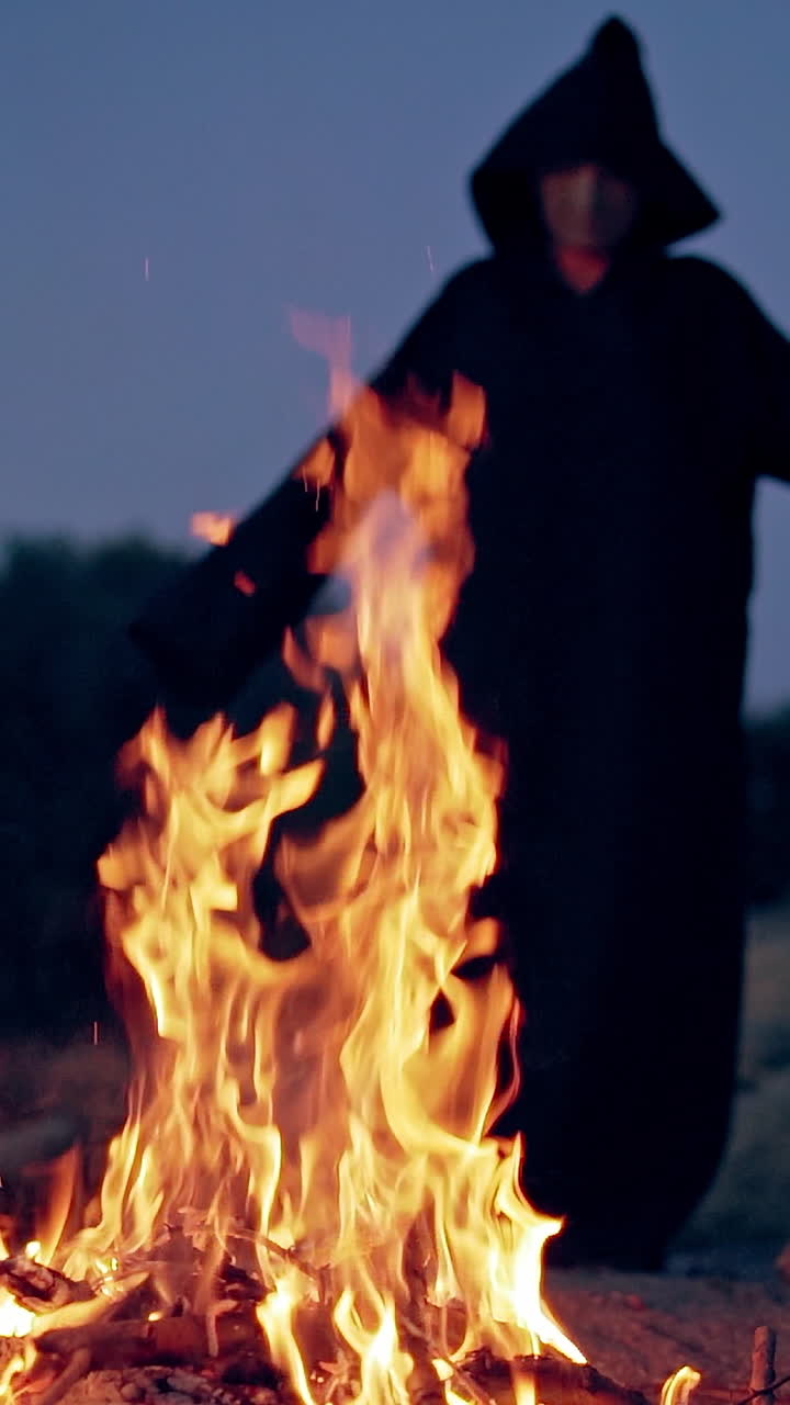 Fire and black figure at darkness. Spooky death in cloak with hood standing against the bright flame and doing some ritual outdoors. Halloween. Vertical video