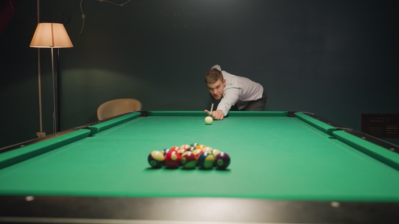 Young man in light shirt and dark trousers leans over green pool table, eyes locked on balls. His posture shows deep concentration as he steadies himself, gripping cue stick. Dim lighting