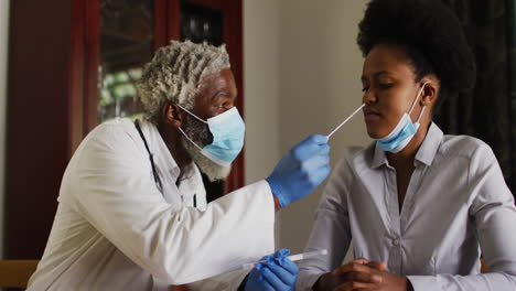 african american senior male doctor wearing face mask swab testing woman for covid at home
