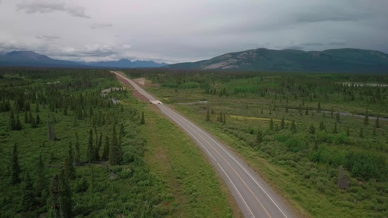 Sensational landscape view of car driving on straight and narrow Alaska highway by Jarvis Creek bridge with sporadic evergreen trees in forest and green grass, America, above aerial static