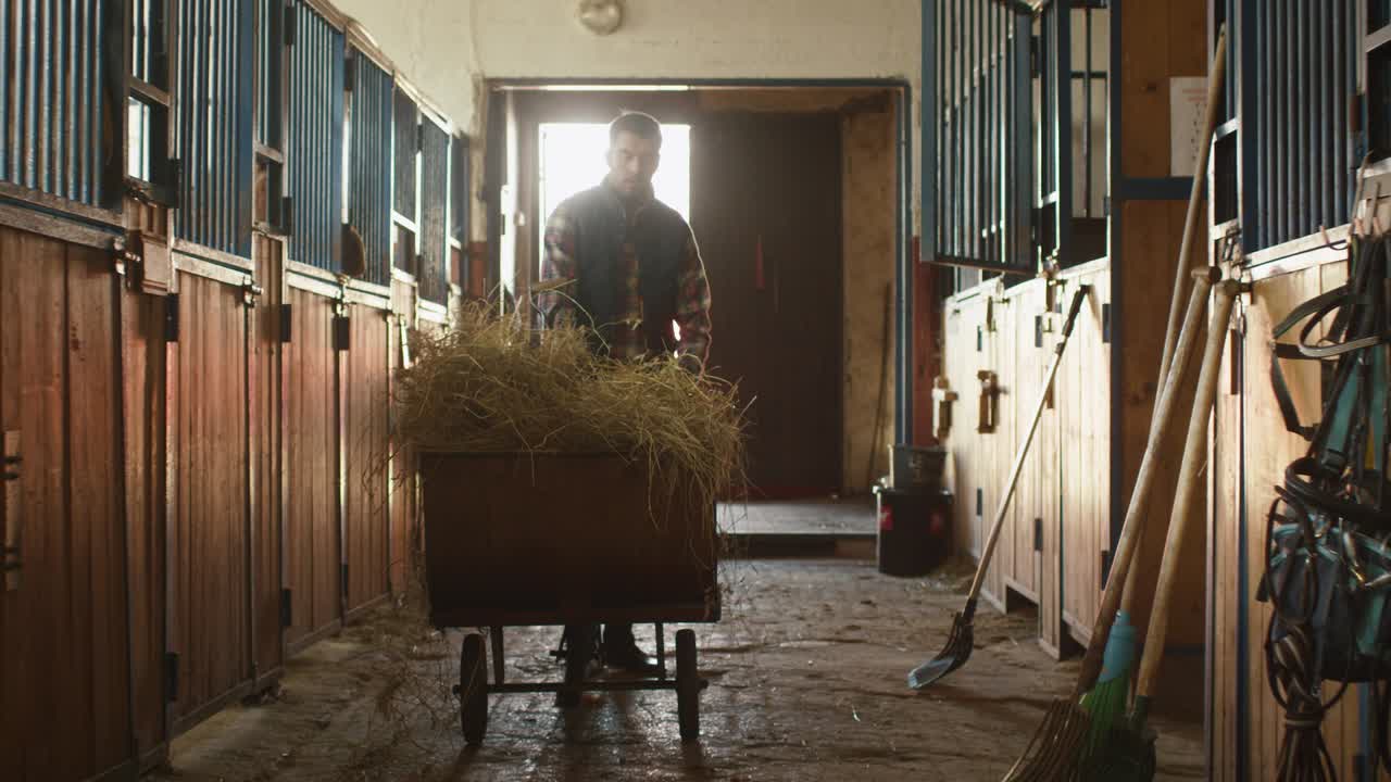 Man is putting a pitchfork back into a cart with hay and moves away from a stable.
