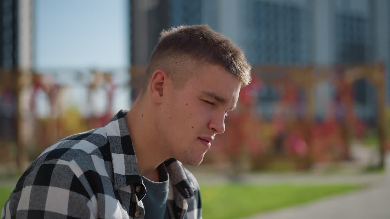 Side view of white man in plaid shirt taking medicine under bright sunlight while seated outdoors, with blurred modern tall office building and colorful garden foliage in background