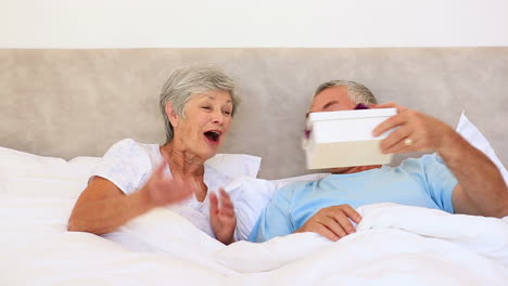 Feliz-Pareja-De-Ancianos-Acostada-En-La-Cama-Con-Un-Hombre-Dando-Un-Regalo