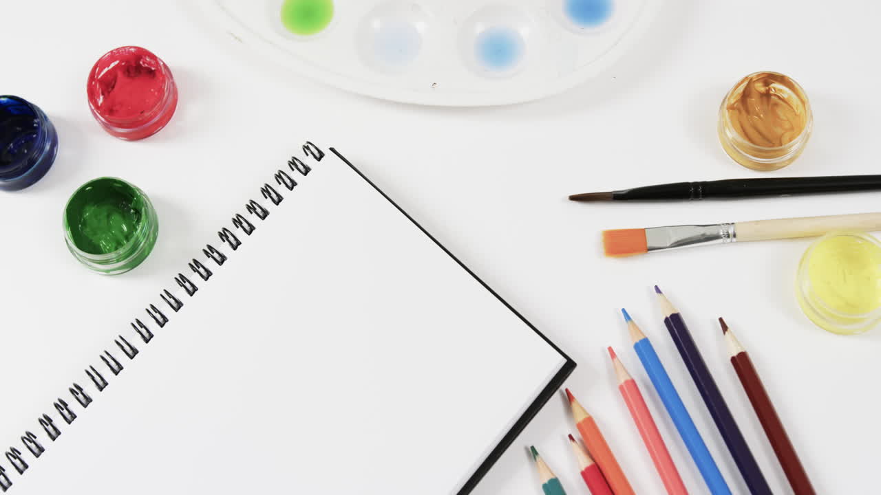 primer plano de un cuaderno, pinturas, pinceles y lápices de colores sobre un fondo blanco, en cámara lenta