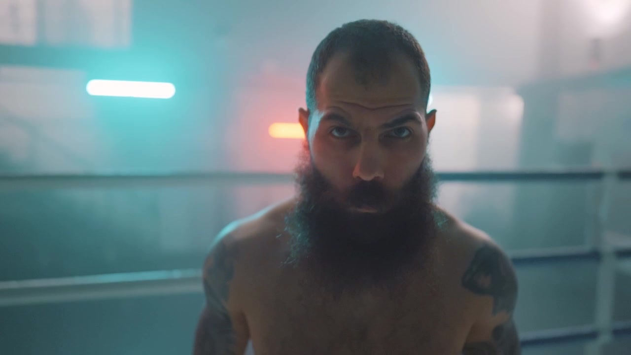 Pugile barbuto dallo sguardo intenso fissa la telecamera in un ring fumoso con luci al neon