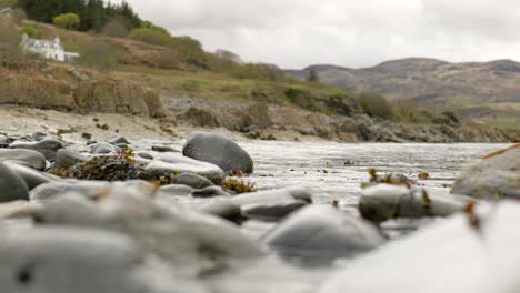 A-low,-sea-level-view-out-to-sea-as-waves-gently-lap-over-wet-boulders-towards-the-camera-with-an-isolated-house-set-against-a-backdrop-of-Scottish-hills