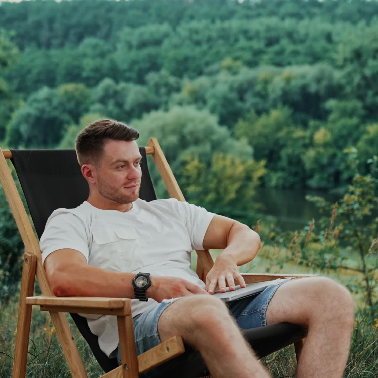 Happy man in a white t-shirt takes a break from work on his laptop. Man puts hands behind his head and relaxes in a chair