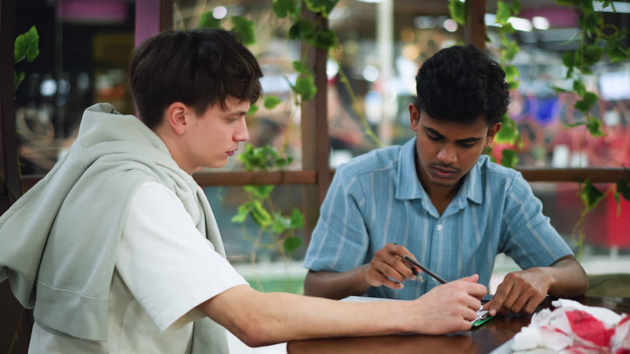 Two friends leaning heads together over wooden desk sketching detailed pencil illustration on white paper with expressions and collaborative energy emphasizing creative partnership and concentration