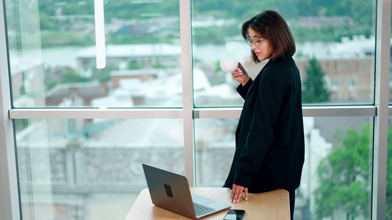 Woman with a cup in hand stands at her desk looking at window. Lady turns to the laptop screen and smiles for a moment.