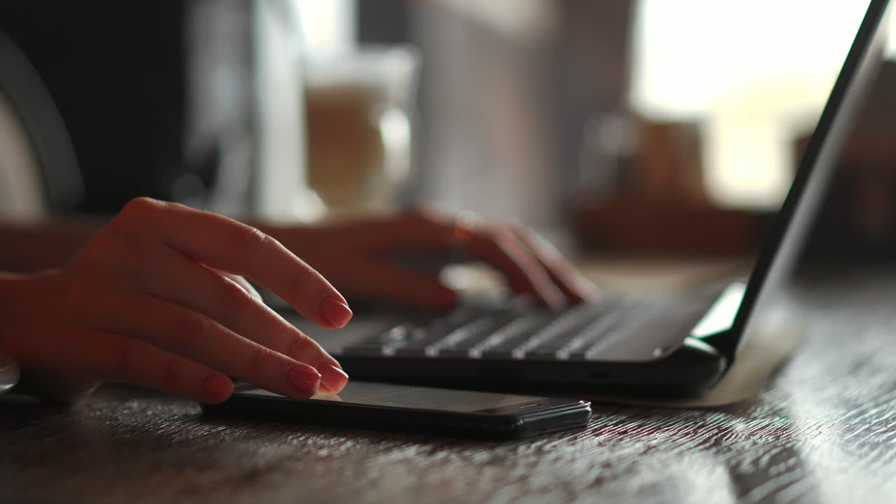 Cute young female adult with grin on phone while working on laptop computer at desk next to coffee cup with large window in background