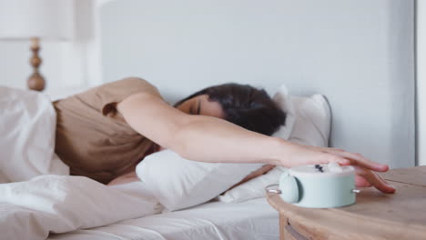 woman asleep in bed reaches out to turn off alarm clock on bedside table