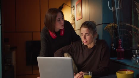 A-brunette-girl-and-a-blonde-girl-have-fun-and-look-at-the-screen-of-a-white-laptop-in-a-dark-room,-with-a-sacred-yellow-lamp,-in-the-evening