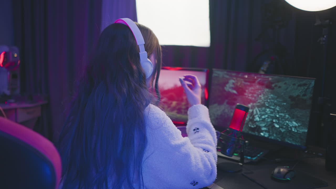 Back view of woman wearing headphones playing video game on dual monitor setup while eating popcorn, dusting hand, and continuing interaction at desk with colorful lighting and streaming microphone