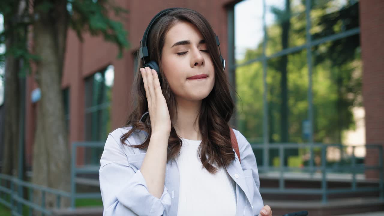 mujer estudiante caucásica usando auriculares y escuchando la música en el teléfono inteligente mientras camina por la calle