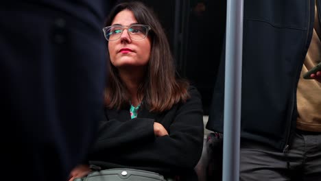 woman with glasses sits on a london underground train in motion, surrounded by other passengers
