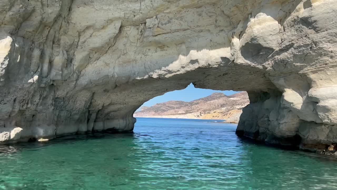 Vibrant cliffs in white, pink, yellow, and red rise above calm turquoise waters of the Aegean Sea under a bright blue sky on Milos Island, Greece.