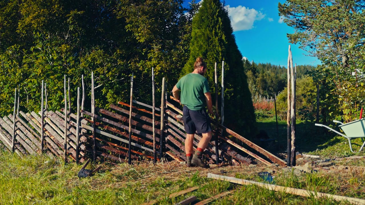A Man is Repairing a Wooden Fence in a Sunny Rural Area, Arranging the Logs and Posts Along the Grassy Field - Static Shot