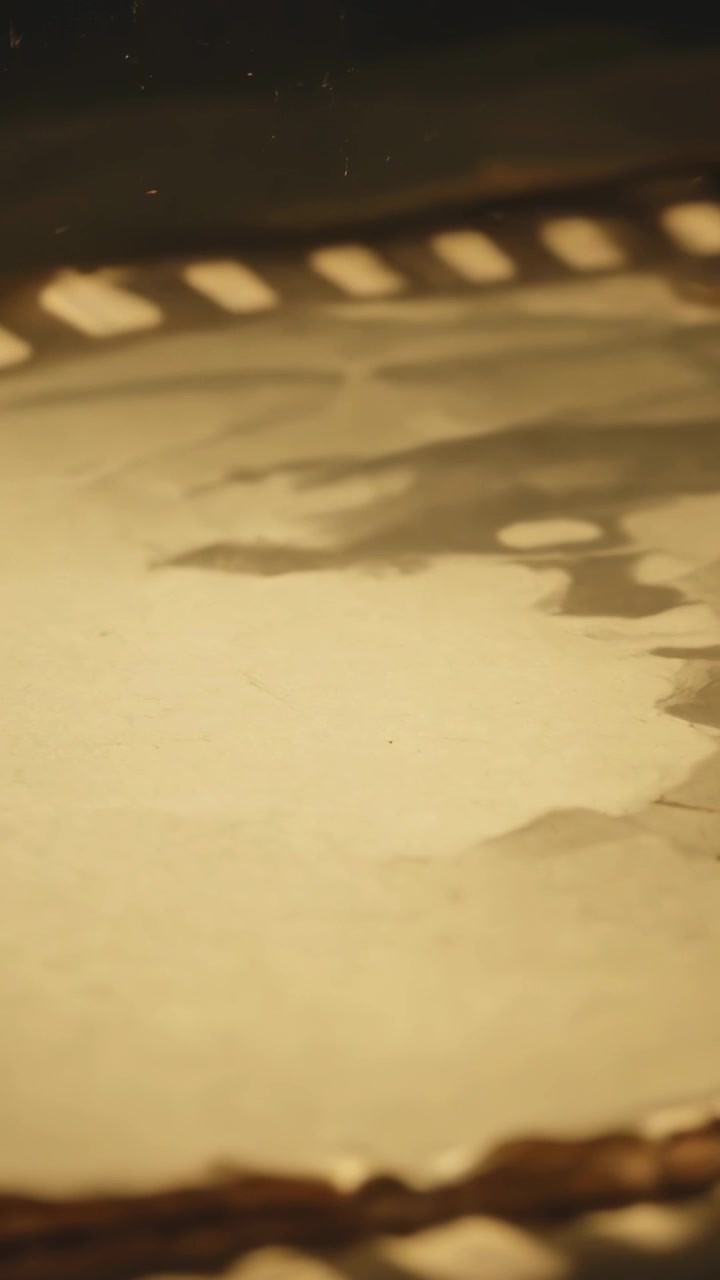 Video verticale: Telecamera in movimento che riprende una pellicola invecchiata in un laboratorio in penombra, ispezionando perforazioni e polvere.