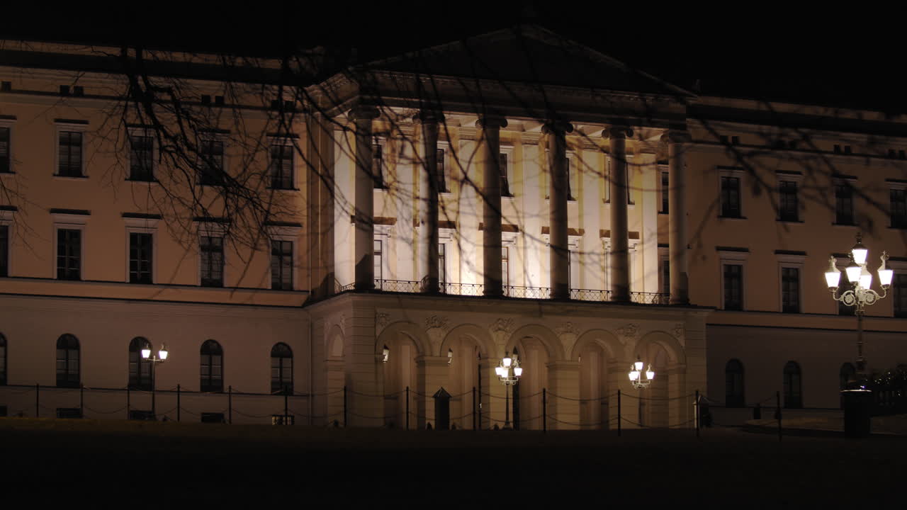 Stabilized slow motion wide 4K shot with parallax motion of tree branches in front of illuminated entrance pillars of the Norwegian Royal Palace on top of Karl Johan street, at night in Oslo Norway
