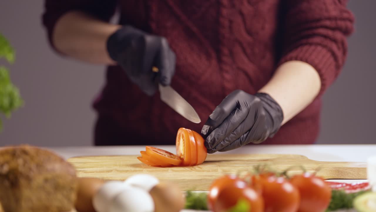A person wearing black gloves slices a ripe red tomato on a wooden board. The fresh, juicy vegetable is being prepped for cooking or a healthy meal, with various ingredients visible in the background