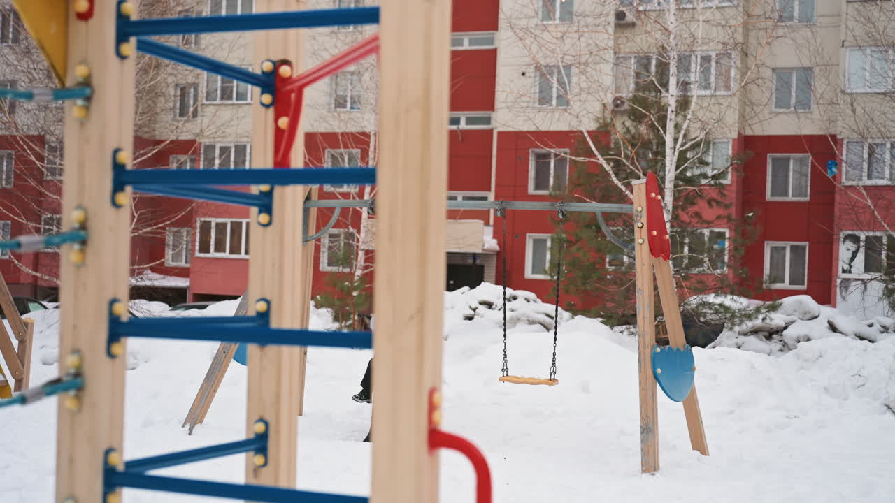 Karlı avluda anne ve oğlu yürüyor, arka planda apartman binası, renkli salıncak seti ve tırmanma parkuru, anne, kaban ve şapka giymiş çocuğunu gözetliyor, taze karda ayak izleri, bulutlu ışık.