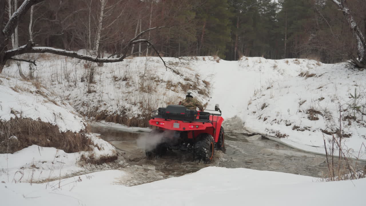ATV rosso che sguazza in un ruscello innevato, pilota in mimetica che attraversa l'acqua bassa. Ampio sfondo di foresta invernale, schizzi di fango e pneumatici chiodati che girano. Azione fuoristrada ad alta energia, estrema e ricca di adrenalina.