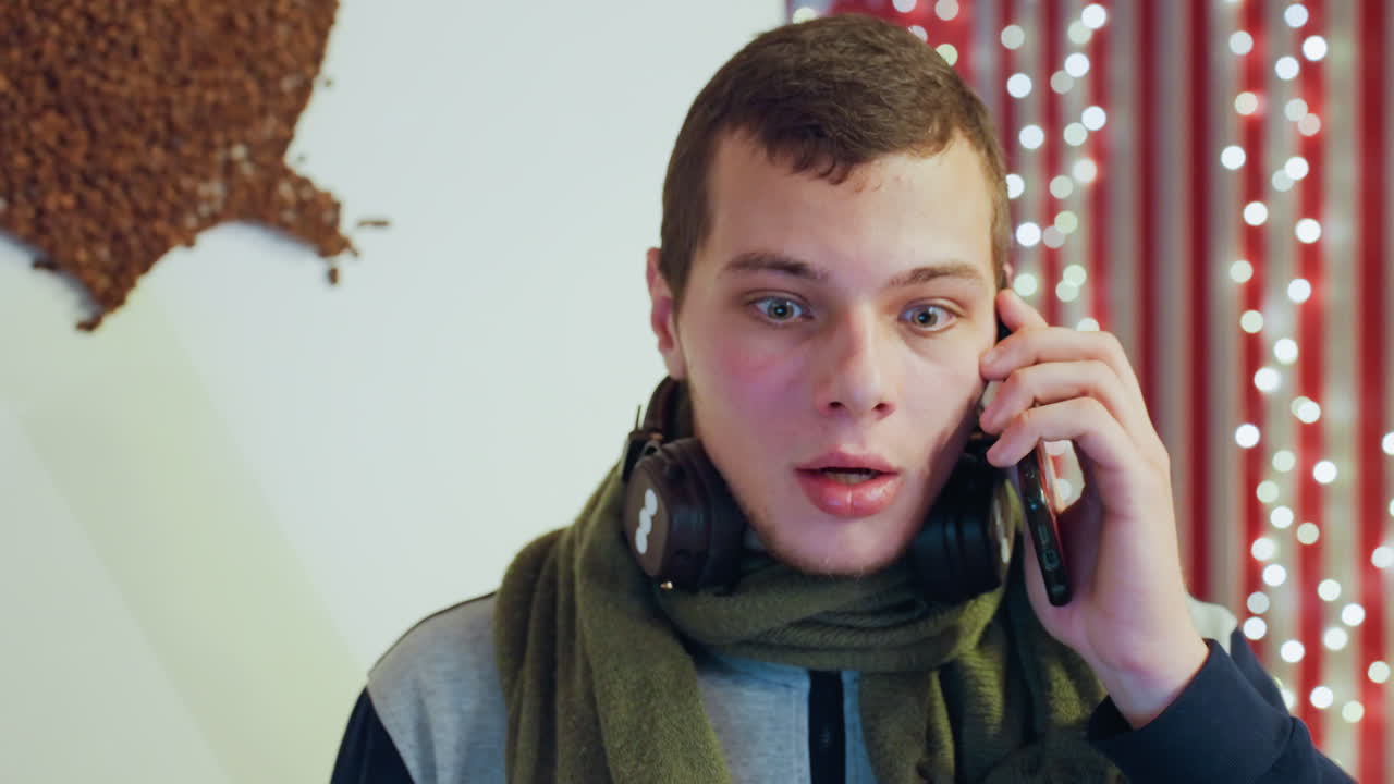 Giovane uomo sorpreso durante una chiamata, tiene lo smartphone mentre sorseggia caffè, cuffie al collo, sfondo di luci festive, occhi espressivi e bocca aperta, scatto spontaneo al chiuso, reazione emotiva.