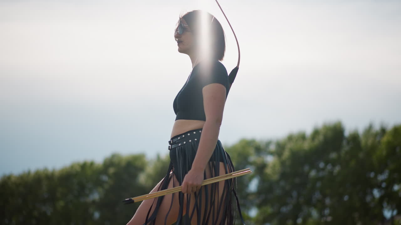 Guerriera solitaria in piedi in un campo, costume di pelle con frange e faretra, che impugna un arco, silhouette di profilo contro un cielo al tramonto incandescente, una leggera brezza muove la gonna, linea di alberi distante a creare