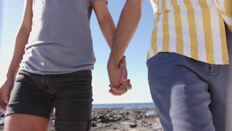 Midsection-of-diverse-gay-male-couple-walking-and-holding-hands-at-beach,-slow-motion