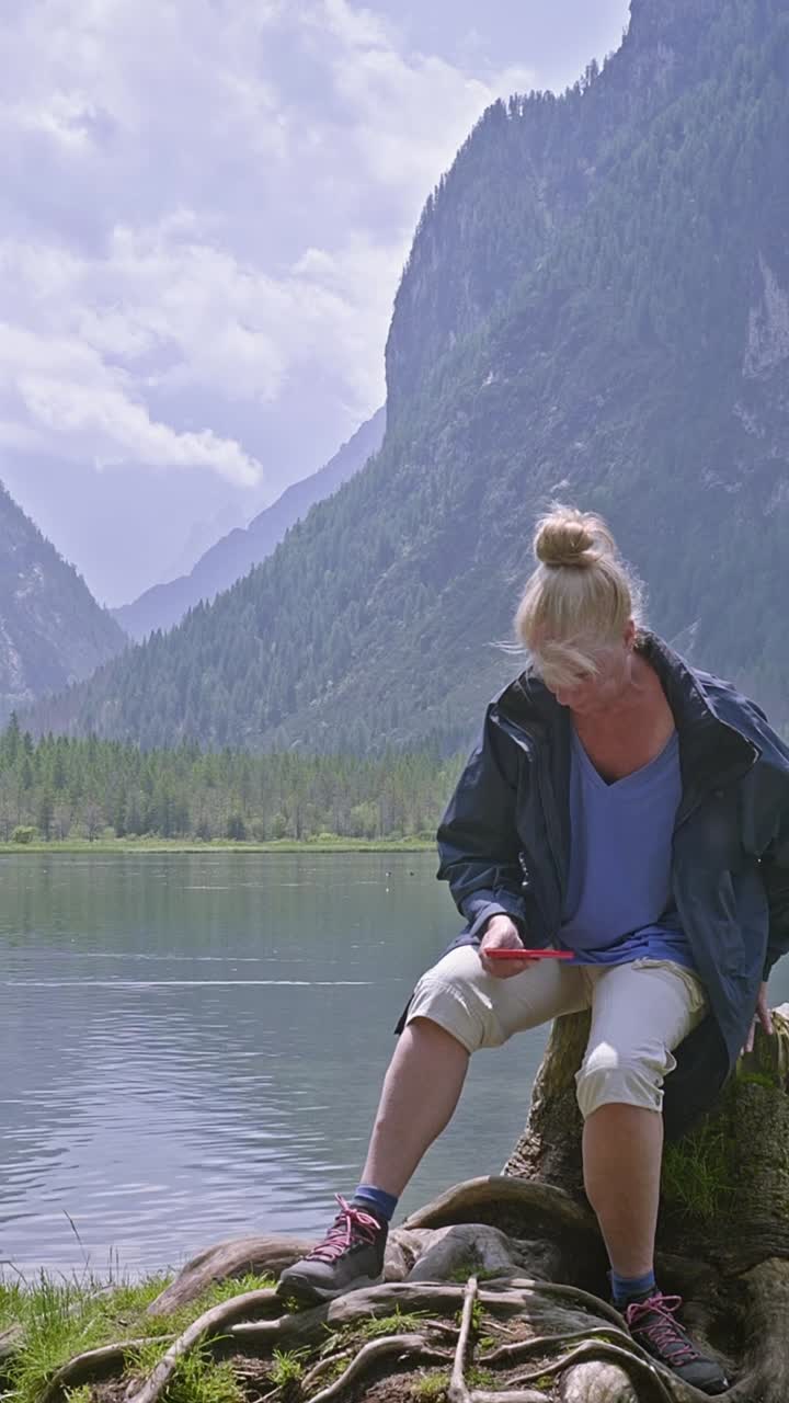Una turista controlla l'app del telefono su un lago panoramico con uno sfondo montuoso.