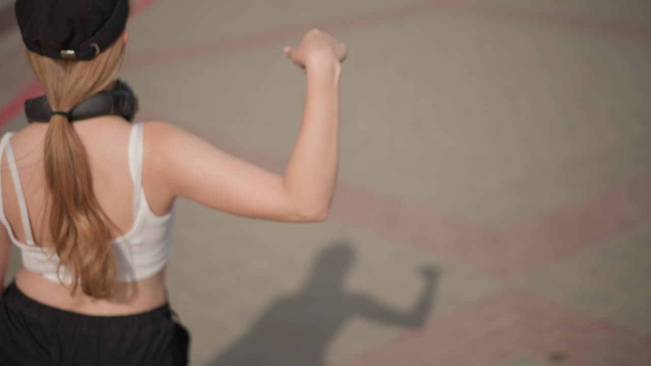 Giovane donna che balla per strada con le cuffie, ripresa da dietro con coda di cavallo e berretto, gesti espressivi con le braccia e schiocchi di dita, ombra sul pavimento decorato, luce solare calda, top corto casual