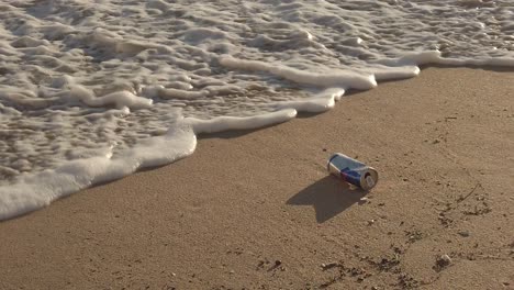 Lata-Vacía-O-Lata-En-La-Playa-De-Arena-Y-Espuma-De-Agua-Procedente-Del-Mar,-Concepto-De-Contaminación-Ambiental