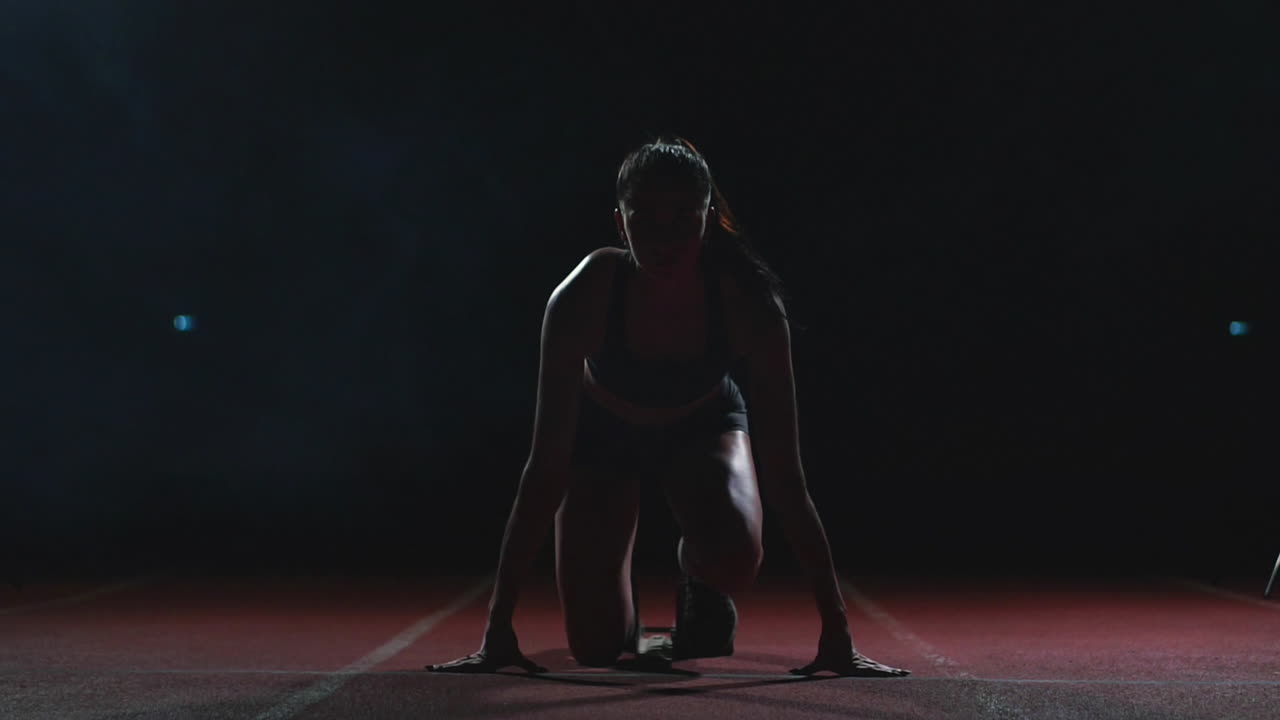 una joven atleta de pantalones cortos negros y una camiseta se está preparando para comenzar en la carrera de 100 metros en la cinta de correr cerca de la línea de salida