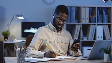 Young-Office-Worker-In-Headphones-Sitting-At-The-Computer-Using-His-Smartphone-And-Writing-In-The-Notebook-While-Looking-At-The-Screen