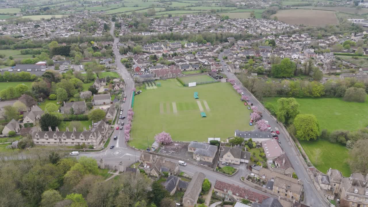 Aerial orbit of manicured cricket ground with vibrant outfield, pavilion and spectators, surrounded by leafy residential streets and blooming trees in a quintessential English setting