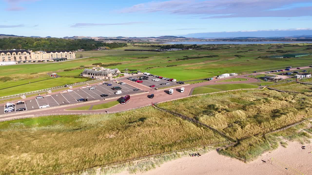 Drone footage glides above sandy beach, grassy dunes, parking area, and historic Victorian architecture near a famous golf course in St Andrews, Scotland. Bright daylight, smooth camera movement